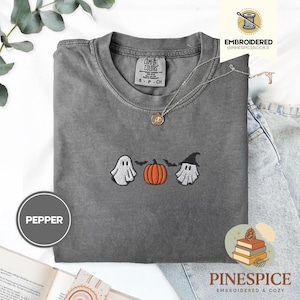 Könnte beinhalten: Ein graues Comfort Colors T-Shirt mit gestickten Halloween-Designs. Das Design umfasst einen weißen Geist, einen orangefarbenen Kürbis und einen Geist mit Hexenhut. Eine silberne Halskette mit Anhänger befindet sich auf dem T-Shirt. Das T-Shirt ist mit "Pepper" gekennzeichnet.