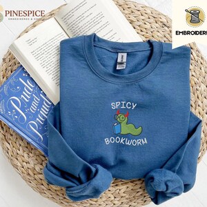 Puede incluir: Una sudadera azul marino con la frase bordada "Spicy Bookworm" y un dise&ntilde;o de gusano de libro de dibujos animados. La sudadera est&aacute; doblada sobre un libro abierto y una copia de "Pride and Prejudice". Los art&iacute;culos est&aacute;n sobre una cesta de mimbre.