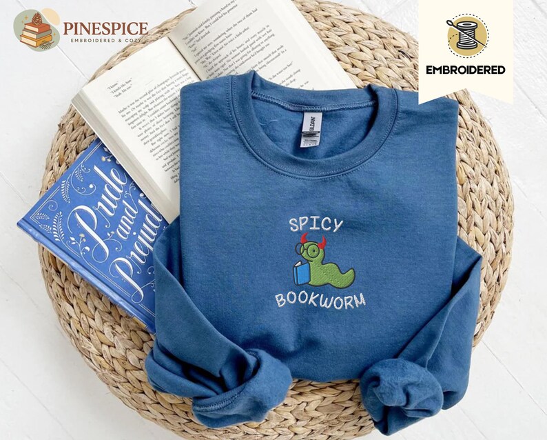 Puede incluir: Una sudadera azul marino con la frase bordada "Spicy Bookworm" y un dise&ntilde;o de gusano de libro de dibujos animados. La sudadera est&aacute; doblada sobre un libro abierto y una copia de "Pride and Prejudice". Los art&iacute;culos est&aacute;n sobre una cesta de mimbre.