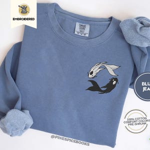 Może przedstawiać: Niebieska bluza dżinsowa z haftowanym wzorem białych i czarnych ryb koi. Bluza ma okrągły dekolt i długie rękawy. Tekst "EMBROIDERED" znajduje się na białej etykiecie. Wykonana w 100% z bawełny.