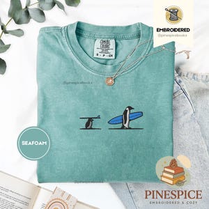 Può includere: T-shirt verde acqua con un disegno ricamato di due pinguini con tavole da surf. Un pinguino porta una tavola da surf, l'altro è in piedi con una tavola blu. La maglietta ha il testo "COMFORT COLORS" e "S - P - CH".