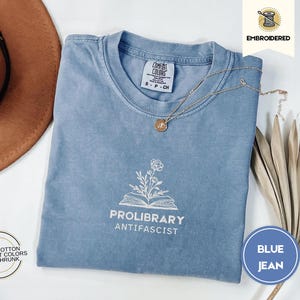 May include: A blue jean coloured t-shirt with the words "PROLIBRARY ANTIFASCIST" printed in white. The shirt has a crew neck and short sleeves. A gold necklace with a pendant is on the shirt. The shirt is made of 100% cotton.