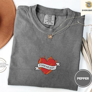 May include: A gray t-shirt with a red embroidered heart with flames and the text "FIREHEART" on a banner. The shirt is made of 100% cotton and is pre-shrunk.