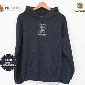 Pu&ograve; includere: Felpa con cappuccio grigio scuro m&eacute;lange con tasca a marsupio e cappuccio con coulisse. La parte anteriore presenta un procione ricamato con le parole "slightly unhinged". La felpa con cappuccio &egrave; esposta su una gruccia di legno.