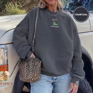 Puede incluir: Sudadera gris carb&oacute;n con el texto "Spicy Bookworm" y un gusano de dibujos animados leyendo un libro. La sudadera tiene cuello redondo y un corte holgado. La modelo lleva vaqueros azules y un bolso con estampado de leopardo.