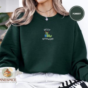 Puede incluir: Una sudadera verde oscuro con la palabra "FOREST" en un c&iacute;rculo. La sudadera tiene un gr&aacute;fico bordado de un gusano de libro verde con un sombrero rojo con las palabras "SPICY BOOKWORM" arriba y abajo del gr&aacute;fico.