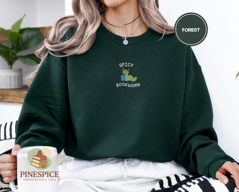 Puede incluir: Una sudadera verde oscuro con la palabra "FOREST" en un c&iacute;rculo. La sudadera tiene un gr&aacute;fico bordado de un gusano de libro verde con un sombrero rojo con las palabras "SPICY BOOKWORM" arriba y abajo del gr&aacute;fico.