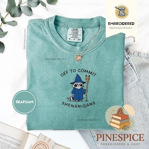 May include: A seafoam green short-sleeve t-shirt featuring an embroidered graphic of a raccoon wearing a blue wizard hat and robe, holding a wooden staff with a skull on top. The text reads OFF TO COMMIT SHENANIGANS in black embroidery.