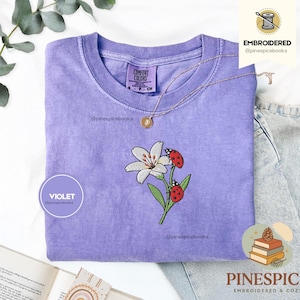 Pode incluir: Uma camiseta violeta com um desenho bordado de um lírio branco e duas joaninhas vermelhas. A camiseta tem gola redonda e mangas curtas. A marca "Comfort Colors" é visível na etiqueta.