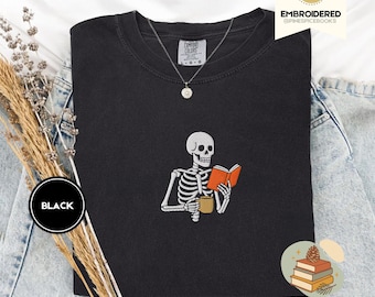 Puede incluir: Sudadera negra con un esqueleto bordado leyendo un libro y sosteniendo una taza. El diseño incluye un collar plateado con un colgante. La sudadera está etiquetada como "Comfort Colors" y "Black". La imagen también incluye el texto "PINESPICE EMBROIDERED & COZY".