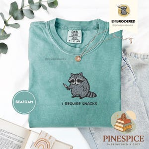 May include: Seafoam green Comfort Colors t-shirt with an embroidered raccoon holding a knife. The text "I REQUIRE SNACKS" is below the raccoon. A silver necklace and a pair of jeans are also visible.