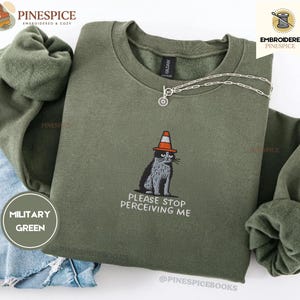 Könnte beinhalten: Ein militärgrüner Rundhals-Pullover mit einer Stickerei, die eine graue Katze mit einem orangefarbenen Verkehrskegel auf dem Kopf zeigt. Unter der Katze steht der Text "PLEASE STOP PERCEIVING ME" in weißen Großbuchstaben.