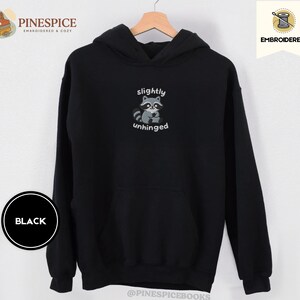 Pu&ograve; includere: Felpa con cappuccio nera con grafica di procione e la scritta "slightly unhinged". Il procione &egrave; grigio e bianco, con il testo in un font semplice. La felpa ha una tasca frontale e un cappuccio.