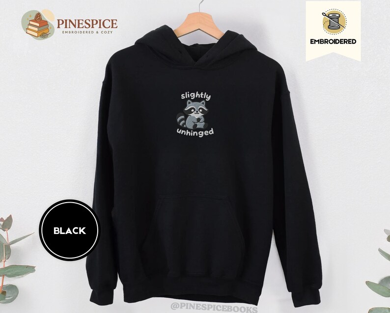 Pu&ograve; includere: Felpa con cappuccio nera con grafica di procione e la scritta "slightly unhinged". Il procione &egrave; grigio e bianco, con il testo in un font semplice. La felpa ha una tasca frontale e un cappuccio.