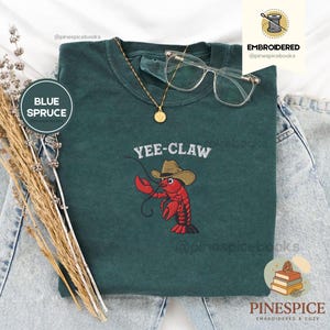 May include: A blue spruce t-shirt with the embroidered text "Yee-Claw" above a red lobster wearing a cowboy hat. The shirt is accessorized with a gold necklace and clear-framed glasses.