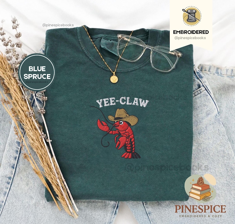 May include: A blue spruce t-shirt with the embroidered text "Yee-Claw" above a red lobster wearing a cowboy hat. The shirt is accessorized with a gold necklace and clear-framed glasses.