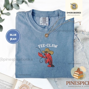 May include: A blue jean Comfort Colors t-shirt with the embroidered phrase "Yee-Claw" above a red lobster wearing a cowboy hat. The shirt has a crew neck and short sleeves. A gold necklace is draped on the shirt.