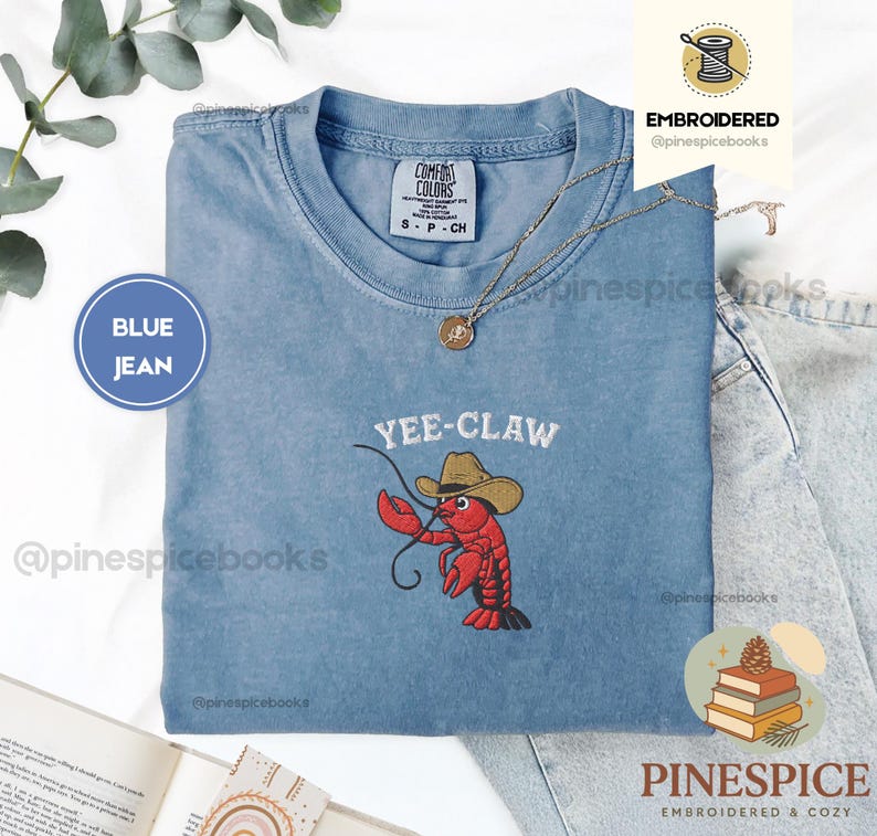 May include: A blue jean Comfort Colors t-shirt with the embroidered phrase "Yee-Claw" above a red lobster wearing a cowboy hat. The shirt has a crew neck and short sleeves. A gold necklace is draped on the shirt.