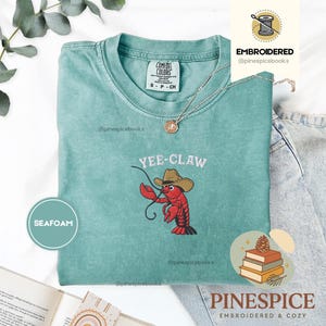 May include: Seafoam green t-shirt with the embroidered phrase "YEE-CLAW" and a cartoon lobster wearing a cowboy hat. The shirt is a Comfort Colors brand, size S-CH. A silver necklace is draped across the shirt.