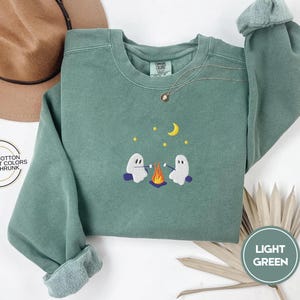 May include: Light green sweatshirt with embroidered design of two ghosts roasting marshmallows over a campfire, with a crescent moon and stars. The sweatshirt has a crew neck and long sleeves. The text "LIGHT GREEN" is in a circle.