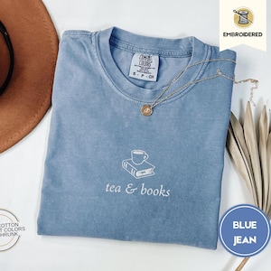 Puede incluir: Una camiseta de mezclilla azul con el texto "tea & books" bordado en la parte delantera. El diseño incluye una taza de té encima de un libro.