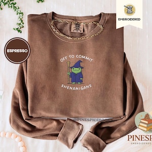 Puede incluir: Sudadera color espresso con el texto "Off to Commit Shenanigans" y una rana de dibujos animados con sombrero y túnica de mago. Una cadena dorada rodea el cuello. La sudadera es bordada y acogedora.