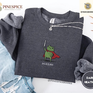 May include: Dark heather gray sweatshirt with an embroidered frog wearing a red cape and holding a sword. The word "Huzzah" is embroidered below the frog. A silver necklace is draped across the sweatshirt. The brand name "Pinespice" is visible.