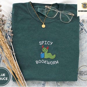 Peut inclure: Un t-shirt vert foncé avec le texte "Spicy Bookworm" brodé en blanc. Le motif représente un ver vert avec des cornes rouges en train de lire un livre.