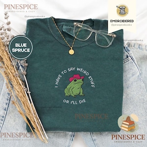 Könnte beinhalten: Ein dunkelgrünes T-Shirt mit einer Stickerei eines grünen Frosches, der einen rosa Cowboyhut trägt. Der Text um den Frosch lautet, I HAVE TO SAY WEIRD STUFF OR I'LL DIE. Eine goldene Halskette mit rundem Anhänger und eine klare Brille liegen darauf.