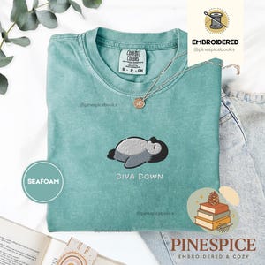 May include: Seafoam green t-shirt with an embroidered design of a sleeping cartoon character and the words "DIVA DOWN". The shirt has a crew neck and short sleeves. A silver necklace with a pendant is on the shirt.