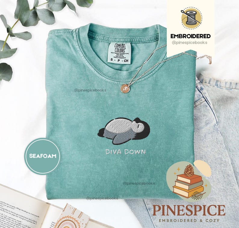 May include: Seafoam green t-shirt with an embroidered design of a sleeping cartoon character and the words "DIVA DOWN". The shirt has a crew neck and short sleeves. A silver necklace with a pendant is on the shirt.