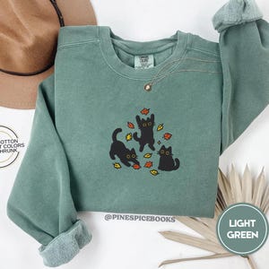 Pode incluir: Camisola verde claro com gola redonda e mangas compridas. O design bordado apresenta três gatos pretos a brincar entre folhas de outono. O texto "@PINESPICEBOOKS" é visível. A etiqueta diz "LIGHT GREEN".