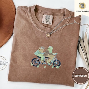 May include: Espresso-colored t-shirt with an embroidered design of two frogs riding a tandem bicycle. The shirt is made of 100% cotton and pre-shrunk. A silver necklace with a pendant rests on the shirt. The tag reads "Comfort Colors".