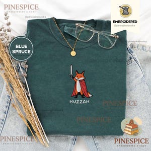 Può includere: Una t-shirt verde scuro con il ricamo di una volpe arancione che indossa un mantello rosso e impugna una spada argentata. Sotto la volpe, il testo riporta la scritta HUZZAH. La t-shirt è abbinata a una collana a catena dorata e occhiali.