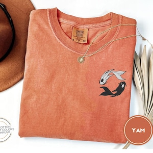 May include: A coral orange t-shirt with a black and white yin yang fish design. The shirt is made of 100% cotton and is pre-shrunk.