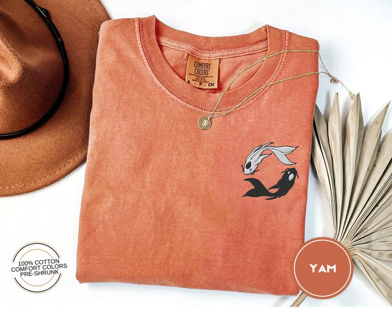 May include: A coral orange t-shirt with a black and white yin yang fish design. The shirt is made of 100% cotton and is pre-shrunk.