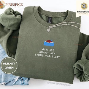 May include: A military green crewneck sweatshirt featuring an embroidered graphic of a blue and red book with the text "ASK ME ABOUT MY LIBBY WAITLIST" in white thread. The garment is displayed with a silver chain necklace and blue denim fabric.