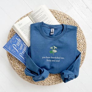 Puede incluir: Una camiseta azul de manga larga con un bordado floral blanco y el texto "you have bewitched me, body and soul".