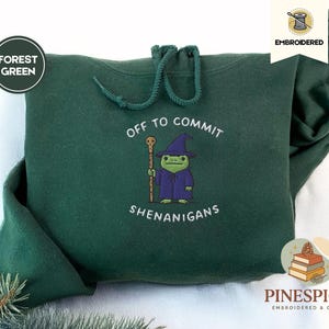 May include: Forest green hoodie with a drawstring hood. The front features an embroidered cartoon frog wizard with the text "OFF TO COMMIT SHENANIGANS". The hoodie has a cozy and comfortable appearance.