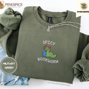 Puede incluir: Sudadera verde militar con la frase bordada "Spicy Bookworm" y un gusano de dibujos animados con gafas y un libro. La sudadera tiene cuello redondo y mangas largas. Un collar plateado est&aacute; en la sudadera.