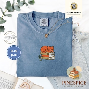 May include: Blue jean colored t-shirt with an embroidered orange cat sleeping on a stack of books. The shirt has the text "COMFORT COLORS" and "BLUE JEAN". A gold necklace with a pendant is on the shirt. The shirt is made of 100% cotton.