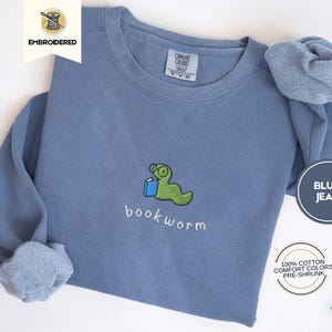May include: Blue jean sweatshirt with an embroidered green bookworm reading a book. The text "bookworm" is embroidered below the image. The sweatshirt is 100% cotton, comfort colors, and pre-shrunk.