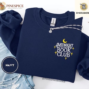 May include: A navy blue sweatshirt with the embroidered text "Midnight Book Club" in white and yellow. The design features a crescent moon and stars. A silver necklace with a circular pendant is on the sweatshirt. The word "NAVY" is in a navy circle.
