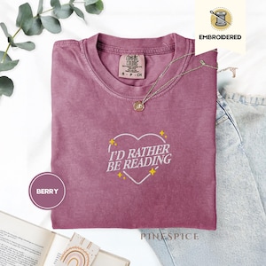 May include: Berry-colored t-shirt with the embroidered phrase "I'D RATHER BE READING" inside a heart shape. The shirt has a crew neck and short sleeves. A gold-colored necklace with a pendant is draped across the shirt. The shirt is made of a soft material.