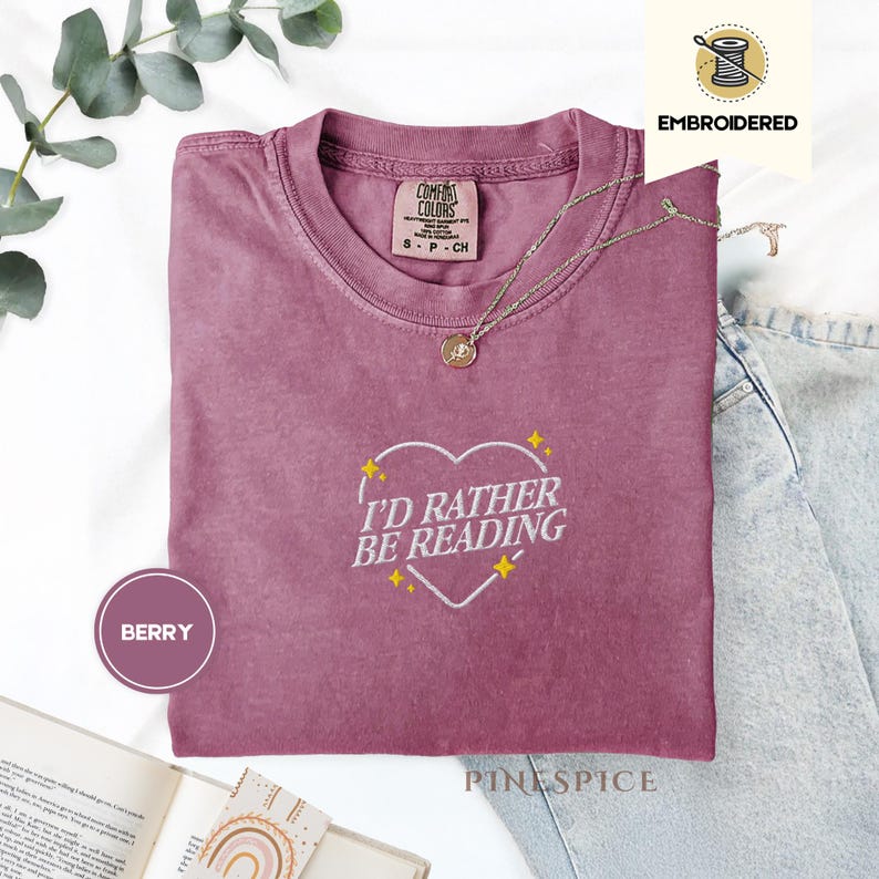 May include: Berry-colored t-shirt with the embroidered phrase "I'D RATHER BE READING" inside a heart shape. The shirt has a crew neck and short sleeves. A gold-colored necklace with a pendant is draped across the shirt. The shirt is made of a soft material.