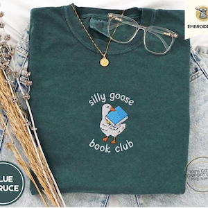 May include: A dark green t-shirt with the text "silly goose book club" embroidered on the front. The text is in white and the design includes a cartoon goose holding a book.