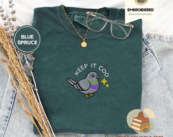 May include: A blue spruce t-shirt with an embroidered pigeon design and the words "Keep it Coo." The shirt also has a gold necklace and clear glasses on top. The brand name "Pinespice" is at the bottom.