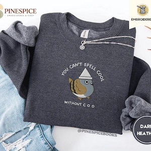 May include: Dark grey sweatshirt featuring an embroidered cartoon bird wearing a hat. The text "You can't spell cool without coo" is embroidered below the bird. A silver necklace is draped across the sweatshirt.