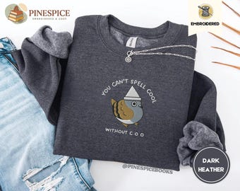 Könnte beinhalten: Dunkelgraues Sweatshirt mit einem gestickten Cartoon-Vogel mit Hut. Der Text "You can't spell cool without coo" ist unter dem Vogel gestickt. Eine silberne Halskette liegt über dem Sweatshirt.