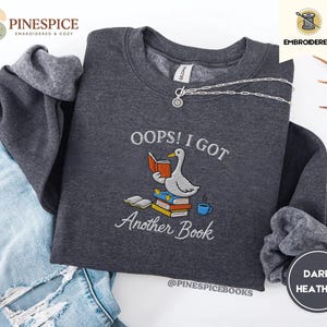 Könnte beinhalten: Ein dunkelgrau meliertes Sweatshirt mit Rundhalsausschnitt und einer Stickerei, die eine weiße Gans zeigt, die auf einem Stapel bunter Bücher sitzt und ein orangefarbenes Buch liest. Der Text lautet OOPS! I GOT Another Book. Eine silberne Kette liegt am Kragen.